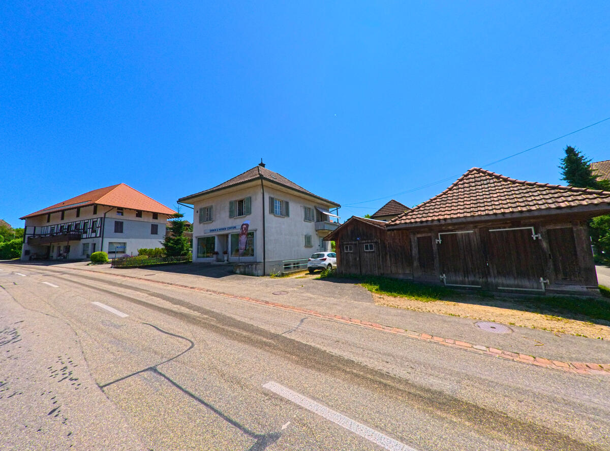 Entlang einer leicht ansteigenden Strasse in Schnottwil (3253, Kanton Solothurn) stehen ein Coiffeursalon, ein Wohnhaus und eine kleine Holzgarage mit Ziegeldach.
