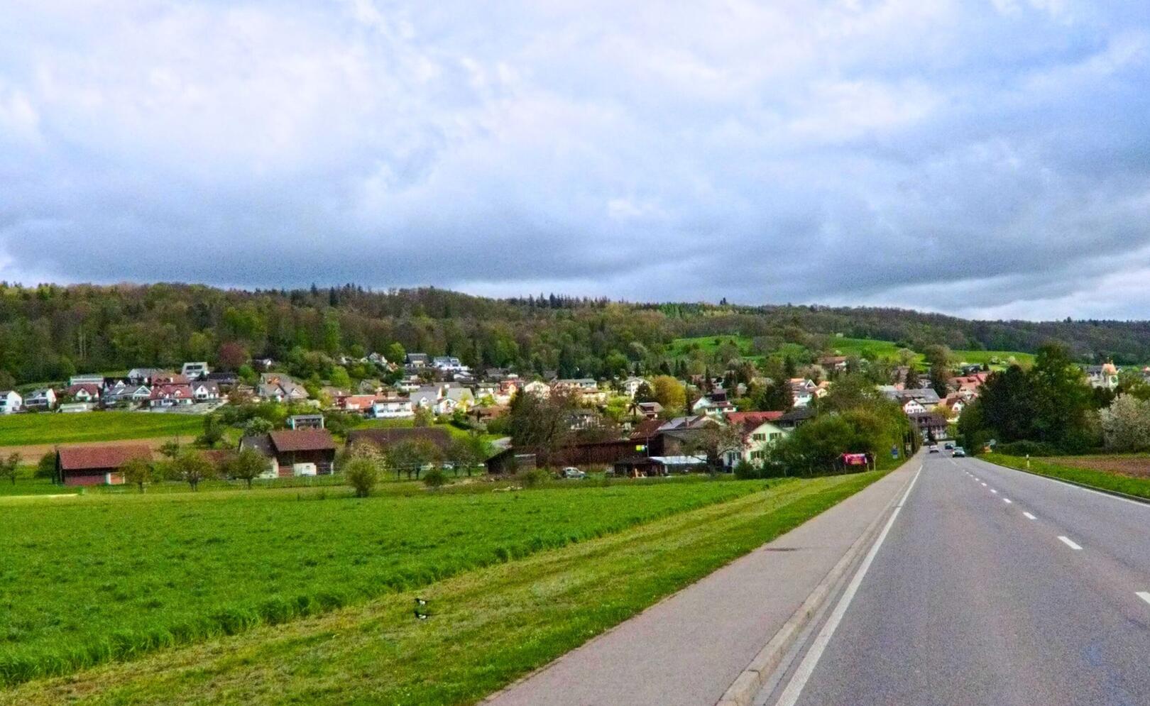 In Schöfflisdorf (8165, Kanton Zürich) führt eine Strasse durch das ländliche Gebiet, wobei im Hintergrund die Wohnhäuser des Ortes sichtbar sind.