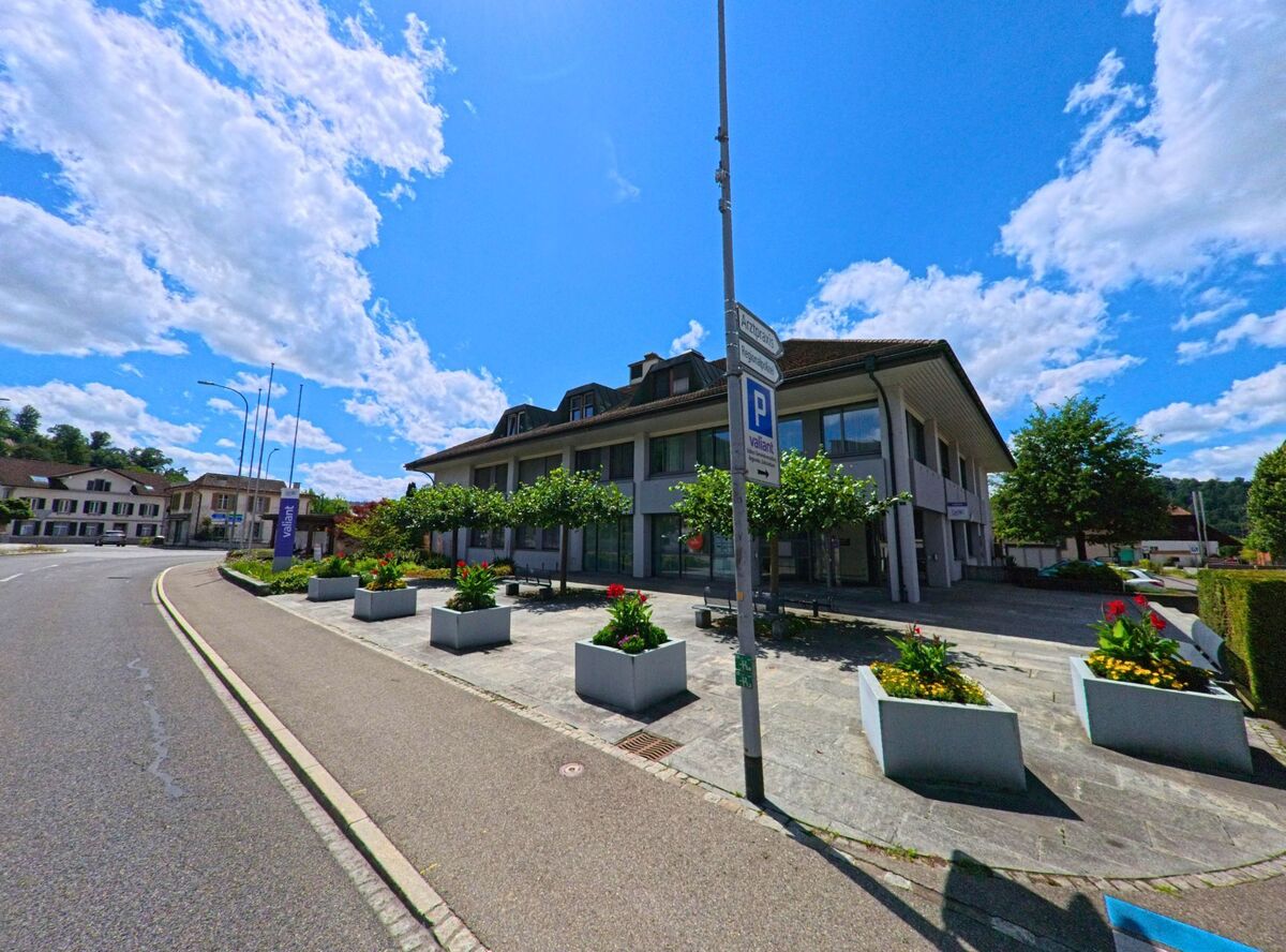 In Schoeftland (5040, Kanton Aargau) steht ein modernes Verwaltungs- und Bankgebaeude mit bepflanztem Vorplatz und Sitzgelegenheiten an einer breiten Strassenkurve.