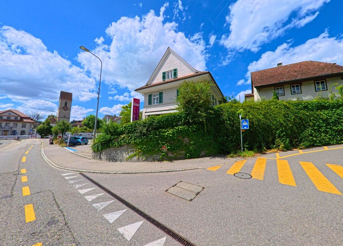 Im Zentrum von Schoeftland (5040, Kanton Aargau) fuehrt eine Strasse mit Fussgaengerstreifen an einem mit Efeu bewachsenen Hang vorbei, im Hintergrund ist der Kirchturm sichtbar.