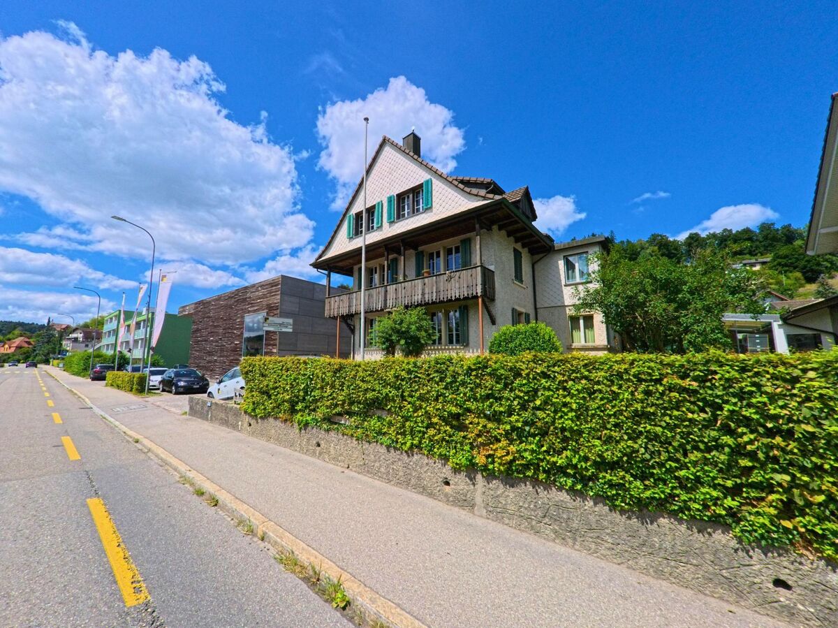 In Schoeftland (5040, Kanton Aargau) steht ein traditionelles Wohnhaus mit gruenen Fensterlaeden und umlaufendem Holzbalkon an einer langen Strasse mit moderner Umgebung.