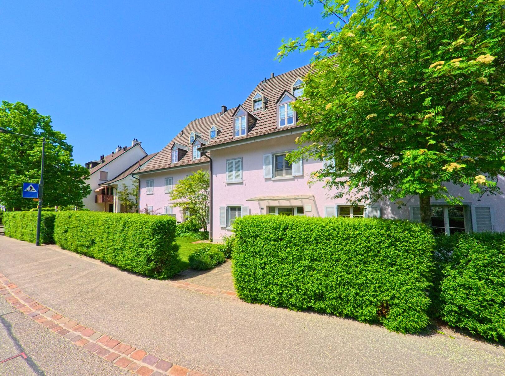 In Schönenbuch (4124, Kanton Baselland) zeigt das Bild ein modernes Wohnhaus mit einem gepflegten Garten und grünen Hecken entlang einer ruhigen Strasse.