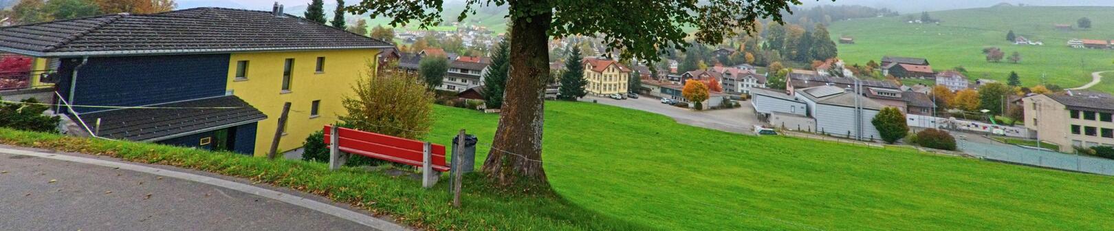 Von einer roten Bank aus in Schönengrund (9105, Kanton Appenzell Ausserrhoden) hat man einen weiten Blick auf die Häuser und grünen Wiesen des Dorfes.