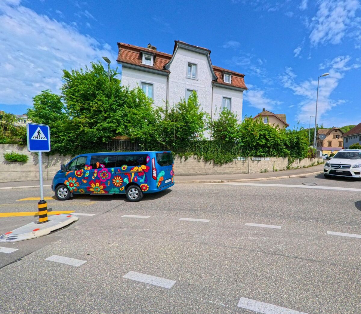 Ein bunt bemalter Kleinbus fährt durch Schönenwerd (5012, Kanton Solothurn) an einem Fussgängerstreifen vorbei, im Hintergrund stehen Wohnhäuser mit Gärten.