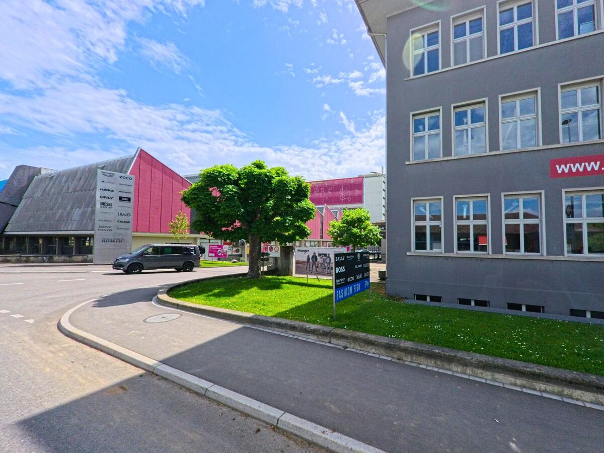 Vor den Gebäuden des Fashion Fish Outlets in Schönenwerd (5012, Kanton Solothurn) stehen Bäume und Wegweiser, die den Eingangsbereich markieren.