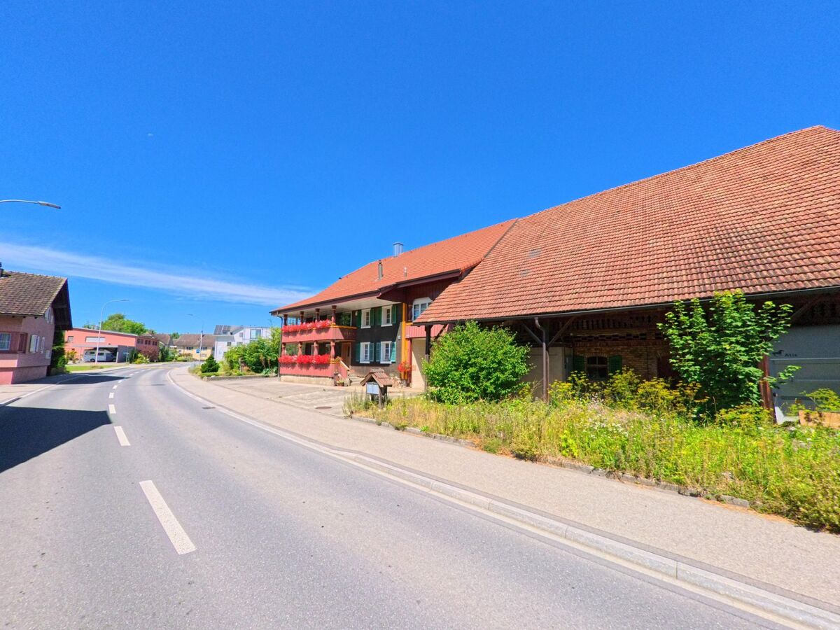 Ein traditionelles Bauernhaus mit roten Fensterladen und angrenzender Scheune steht in Schoetz (6247, Kanton Luzern) an einer sanft geschwungenen Strasse mit Gehweg.