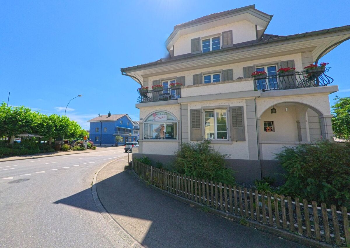 Ein Eckhaus mit Balkonen und Fensterlaeden in Schoetz (6247, Kanton Luzern) liegt an einer leicht gekruemmten Strasse, gegenüber steht ein markant blau gestrichenes Wohnhaus.