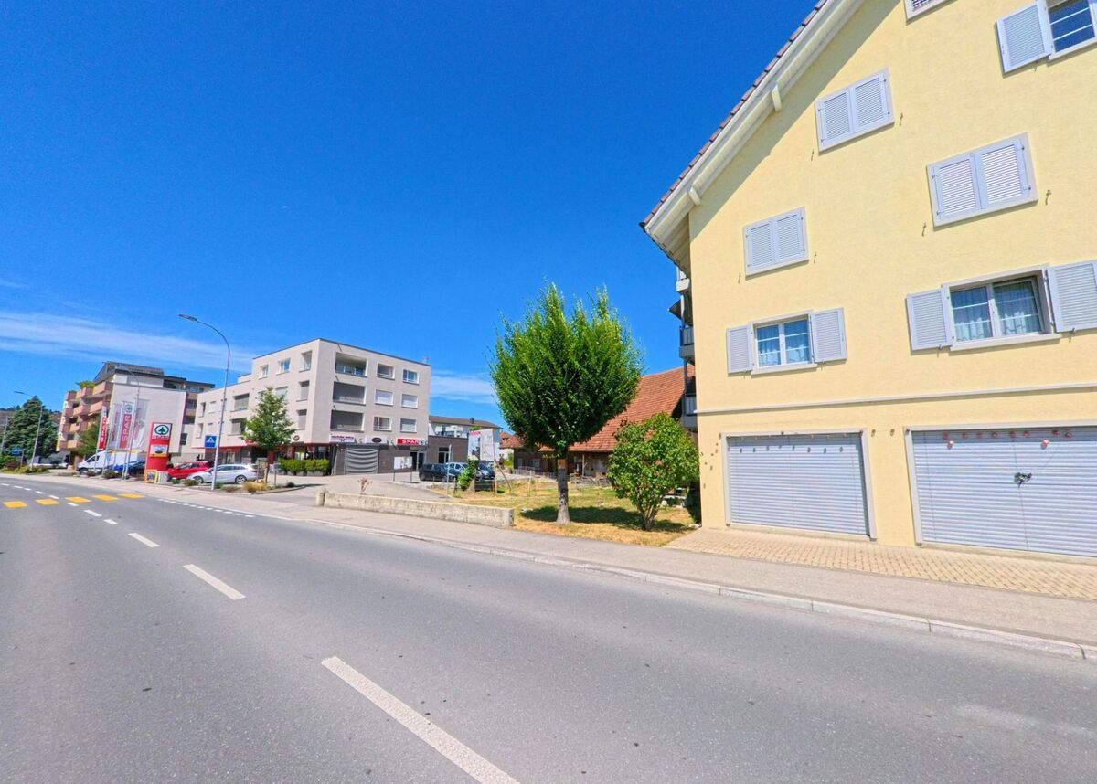 In Schoetz (6247, Kanton Luzern) verläuft eine Strasse mit Fussgaengerstreifen an modernen Wohn- und Geschaeftshaeusern vorbei, links sind ein Autohaendler und weitere Laeden sichtbar.