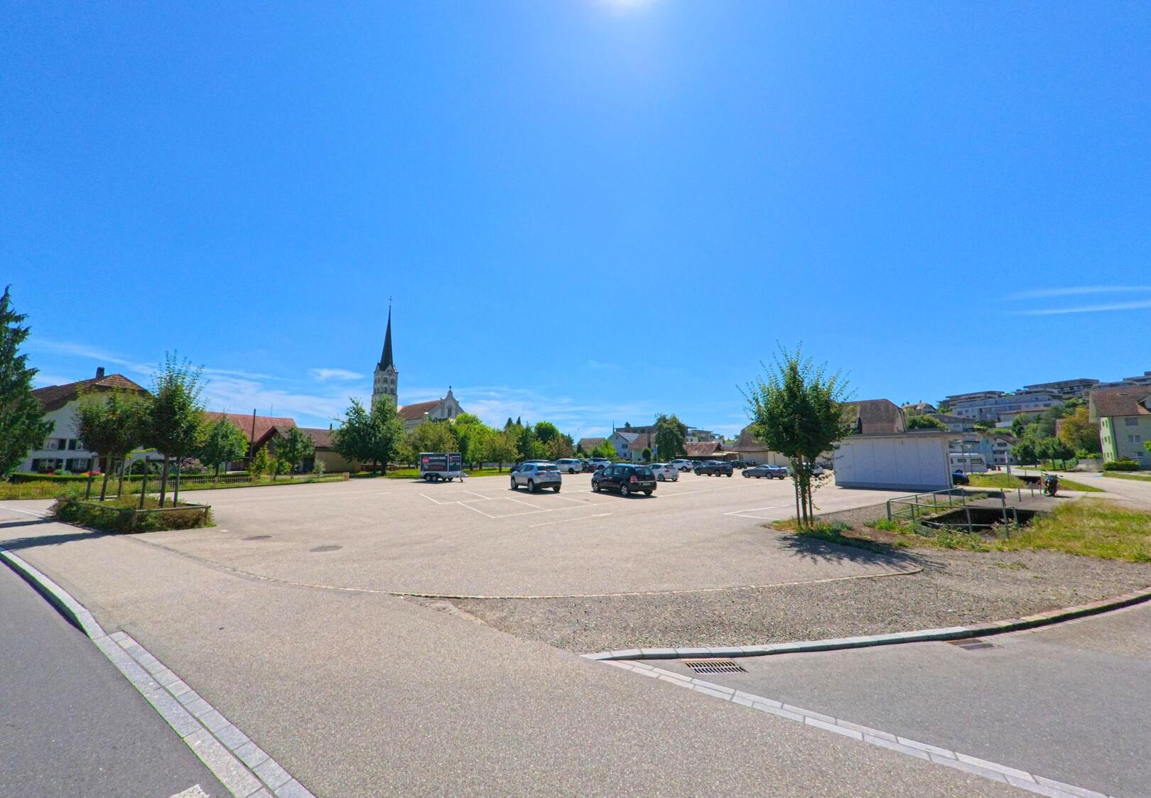 In Schoetz (6247, Kanton Luzern) liegt ein weitlaeufiger Parkplatz unter blauem Himmel, im Hintergrund ragt der Kirchturm über die umliegenden Wohnhaeuser hinweg.