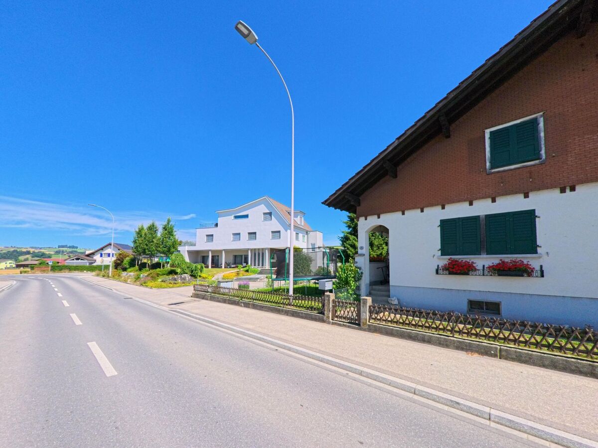 In Schoetz (6247, Kanton Luzern) stehen traditionelle und moderne Wohnhaeuser mit gepflegten Gaerten an einer laendlichen Ausfallstrasse mit Blick auf offene Felder.