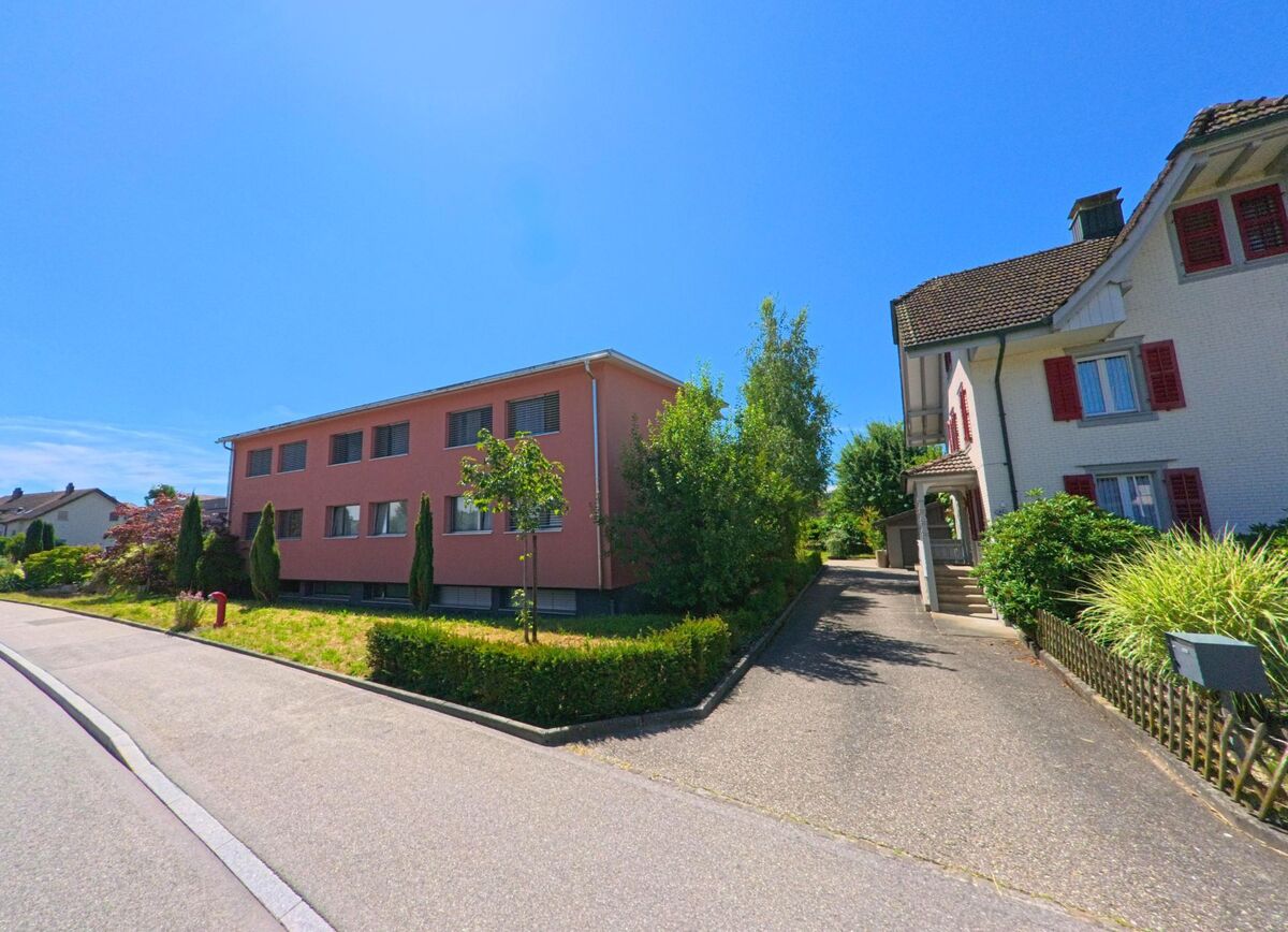 In Schoetz (6247, Kanton Luzern) stehen zwei Wohnhaeuser mit gepflegten Vorgartenanlagen nebeneinander, verbunden durch eine asphaltierte Zufahrt unter strahlend blauem Himmel.