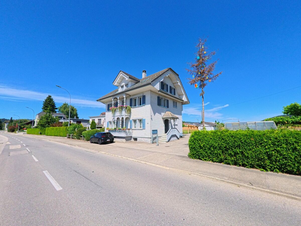 Ein weisses Wohnhaus mit Balkon und verziertem Eingang steht in Schoetz (6247, Kanton Luzern) an einer leicht geschwungenen Strasse, daneben ragt ein geschmueckter Baum in den Himmel.