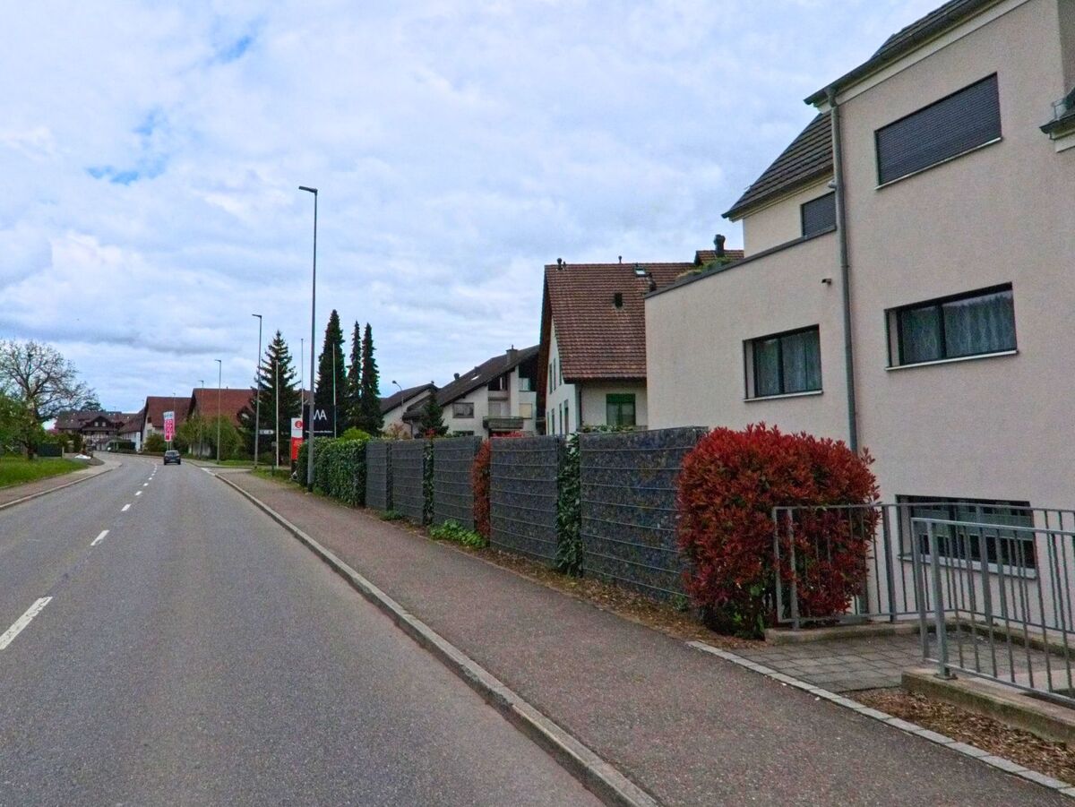 In Schöfflisdorf (8165, Kanton Zürich) verläuft eine Wohnstrasse mit Einfamilienhäusern und gepflegten Vorgärten entlang eines breiten Trottoirs.