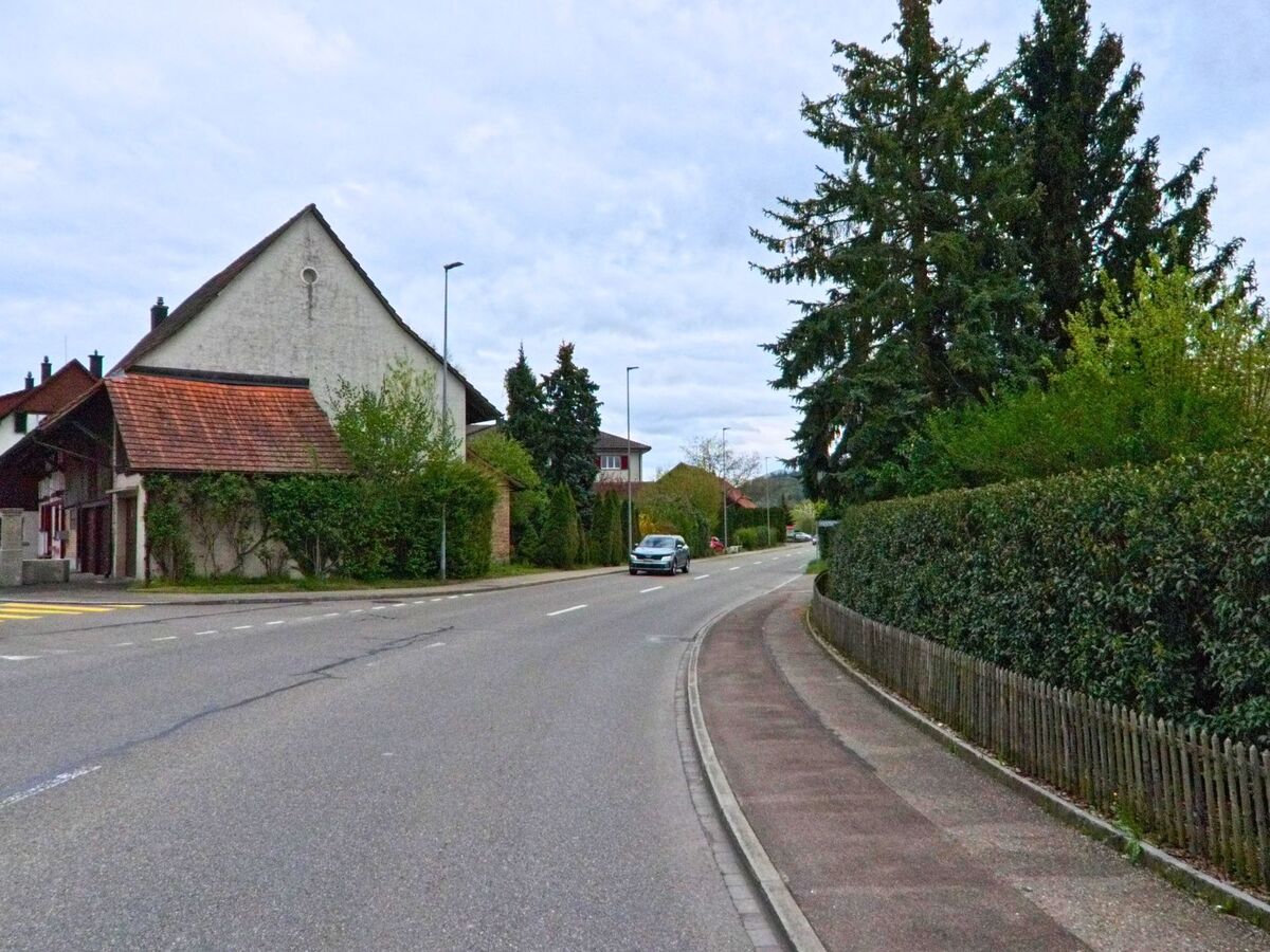 In Schöfflisdorf (8165, Kanton Zürich) windet sich eine Strasse an einem älteren Gebäude mit Ziegeldach und hohen Bäumen entlang durch das Dorf.