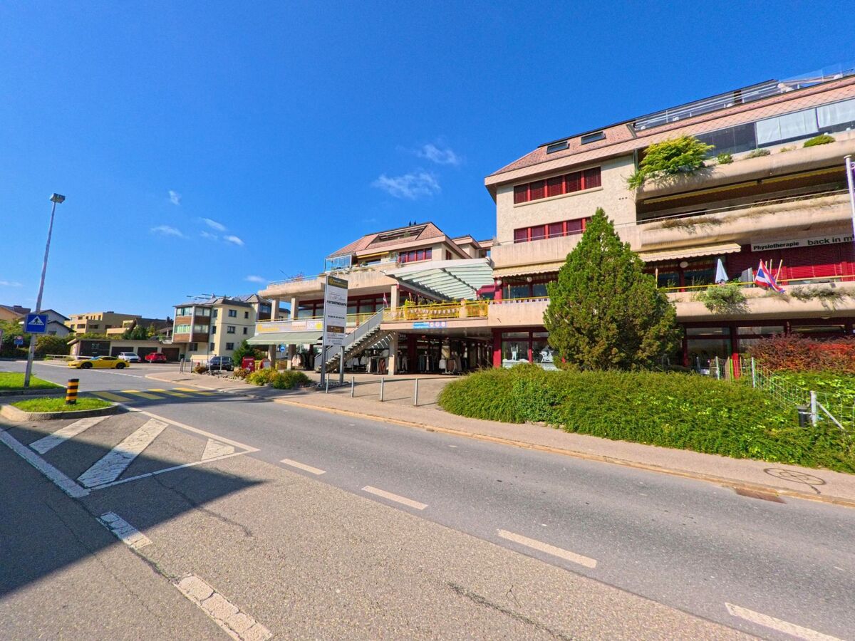 Gegenueber eines mehrstoeckigen Gebaeudekomplexes in Schuebelbach (8862, Kanton Schwyz) verlaeuft eine breite Strasse mit Fussgaengerstreifen und Inselbegruenung.