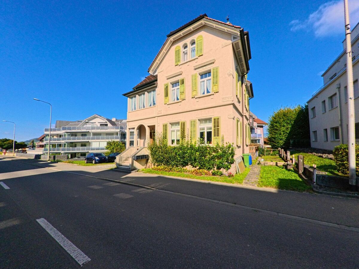 Ein helles Wohnhaus mit gruenen Fensterlaeden steht in Schuebelbach (8862, Kanton Schwyz) an einer Strasse, flankiert von modernen Gebaeuden und gepflegtem Gruen.