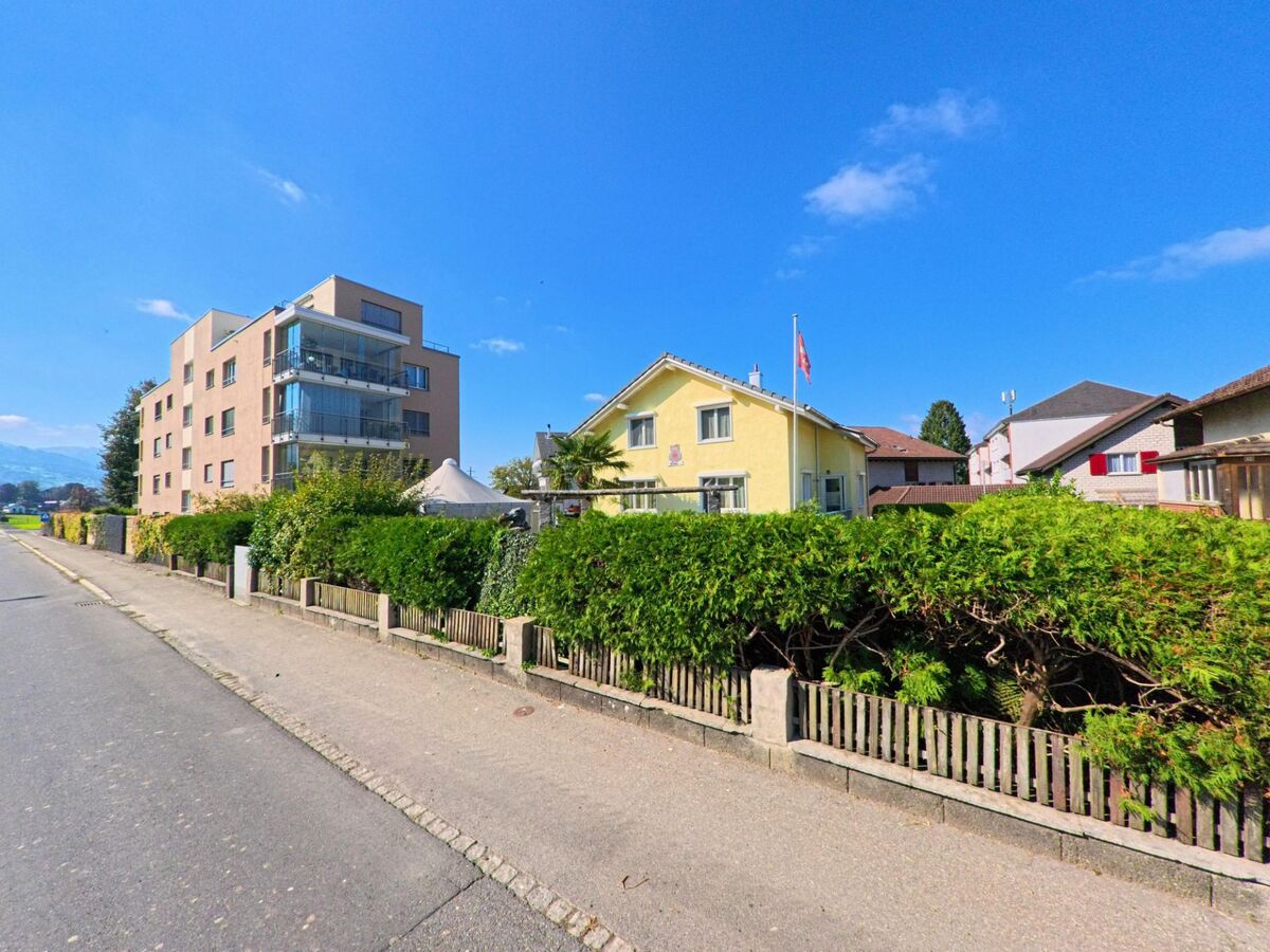 An einer ruhigen Strasse in Schuebelbach (8862, Kanton Schwyz) stehen ein modernes Mehrfamilienhaus und ein gelbes Wohnhaus hinter gepflegten Hecken.