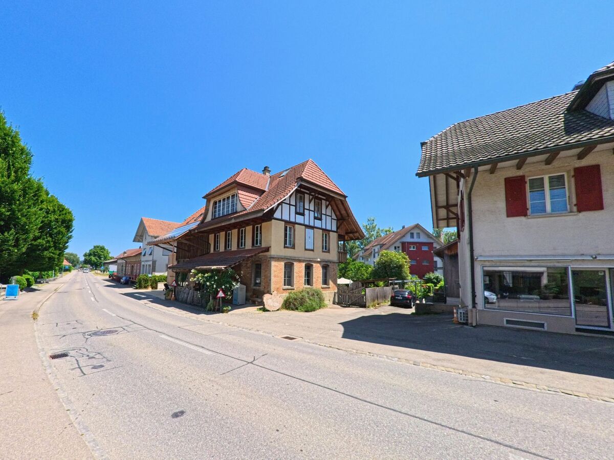 Eine Straße in Schüpfen (3054, Kanton Bern) führt an traditionellen Fachwerkhäusern und modernen Gebäuden entlang, gesäumt von gepflegten Bäumen.