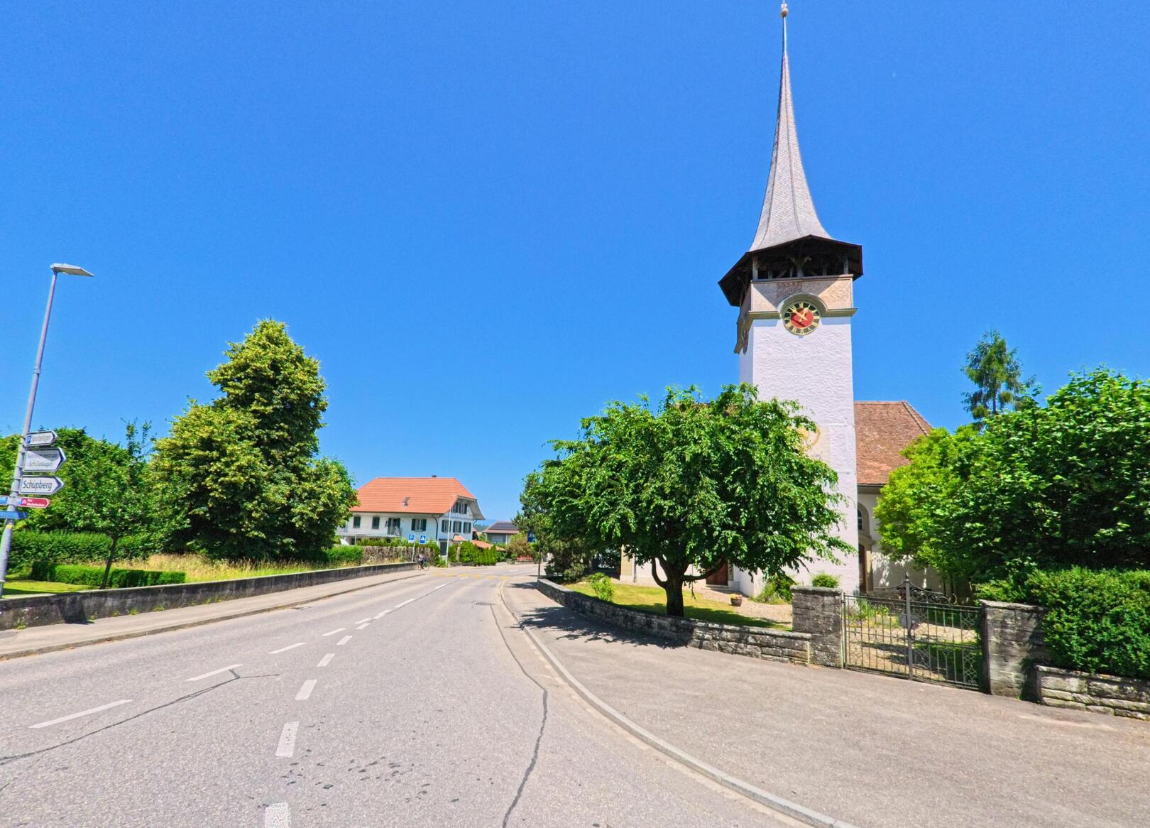 Die Strasse in Schüpfen (3054, Kanton Bern) führt an der imposanten Kirche vorbei und bietet einen klaren Blick auf den Himmel und die umgebenden Gebäude.