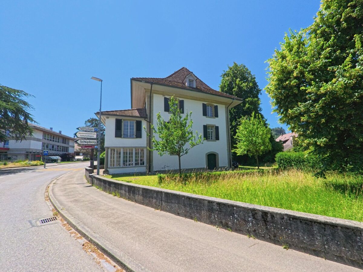 Ein traditionelles Gebäude in Schüpfen (3054, Kanton Bern), umgeben von grünen Wiesen und Bäumen, mit Wegweisern zu den umliegenden Orten.