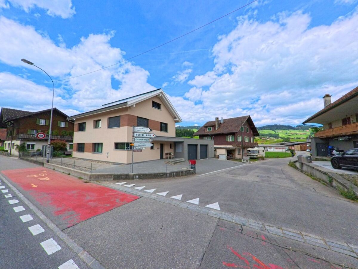 Am Ortseingang von Schüpfheim (6170, Kanton Luzern) markieren Wegweiser und ein modernes Wohnhaus mit Velostreifen den Übergang zur ländlichen Umgebung mit grünen Hügeln.