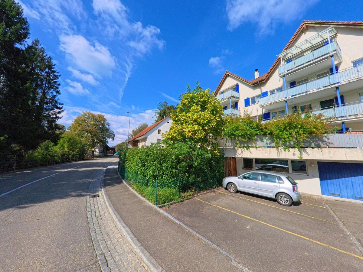 Mehrfamilienhäuser mit Balkonen und blauen Fensterläden stehen in Schupfart (4325, Kanton Aargau) entlang einer leicht geschwungenen Strasse mit angrenzenden Bäumen.