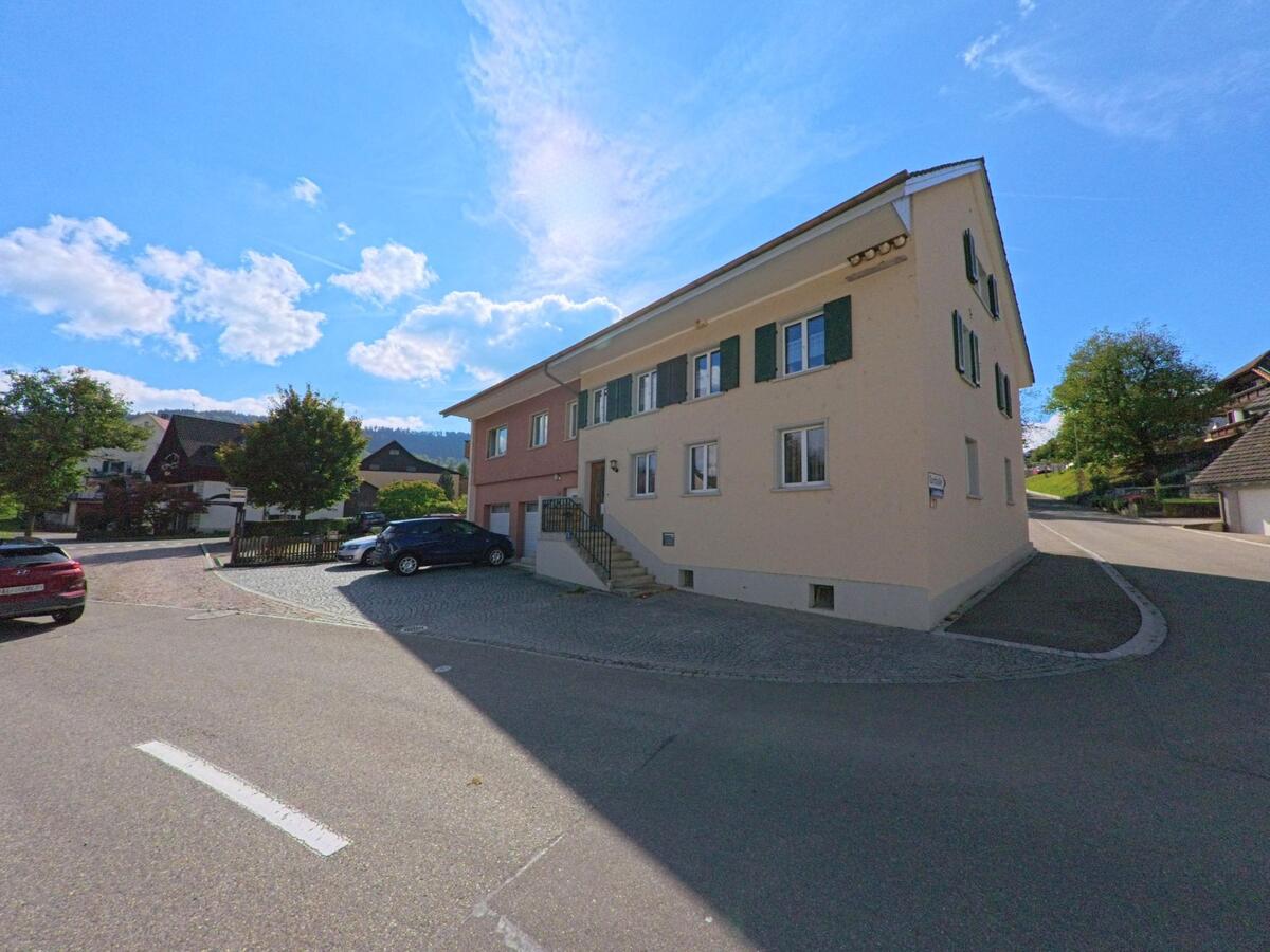 Ein helles Wohnhaus mit grünen Fensterläden steht an einer Strassenecke in Schupfart (4325, Kanton Aargau), umgeben von weiteren Gebäuden und parkenden Autos.