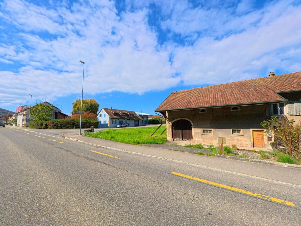 Ein altes Bauernhaus mit grossem Holztor steht an einer breiten Strasse in Schwaderloch (5326, Kanton Aargau) neben einer grünen Wiese unter blauem Himmel.