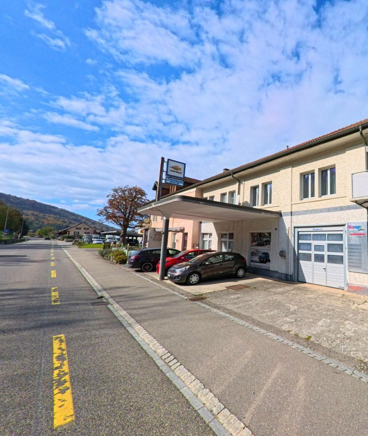 Ein Garagenbetrieb mit ueberdachtem Eingang und geparkten Autos liegt an der Hauptstrasse in Schwaderloch (5326, Kanton Aargau) mit Blick auf die bewaldeten Huegel.