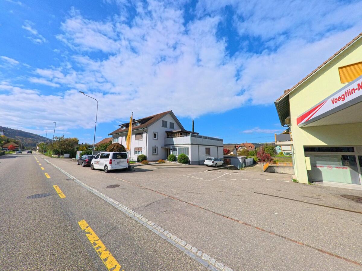Ein Wohn- und Geschaeftsgebaeude mit Parkplatz liegt an der Hauptstrasse von Schwaderloch (5326, Kanton Aargau) bei klarem Wetter und leichtem Verkehr.