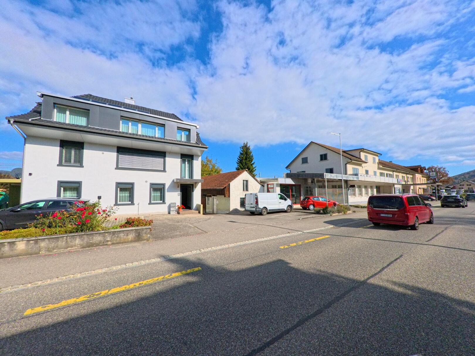Entlang der Hauptstrasse in Schwaderloch (5326, Kanton Aargau) stehen moderne Wohn- und Geschäftsgebäude unter einem leicht bewölkten Himmel.