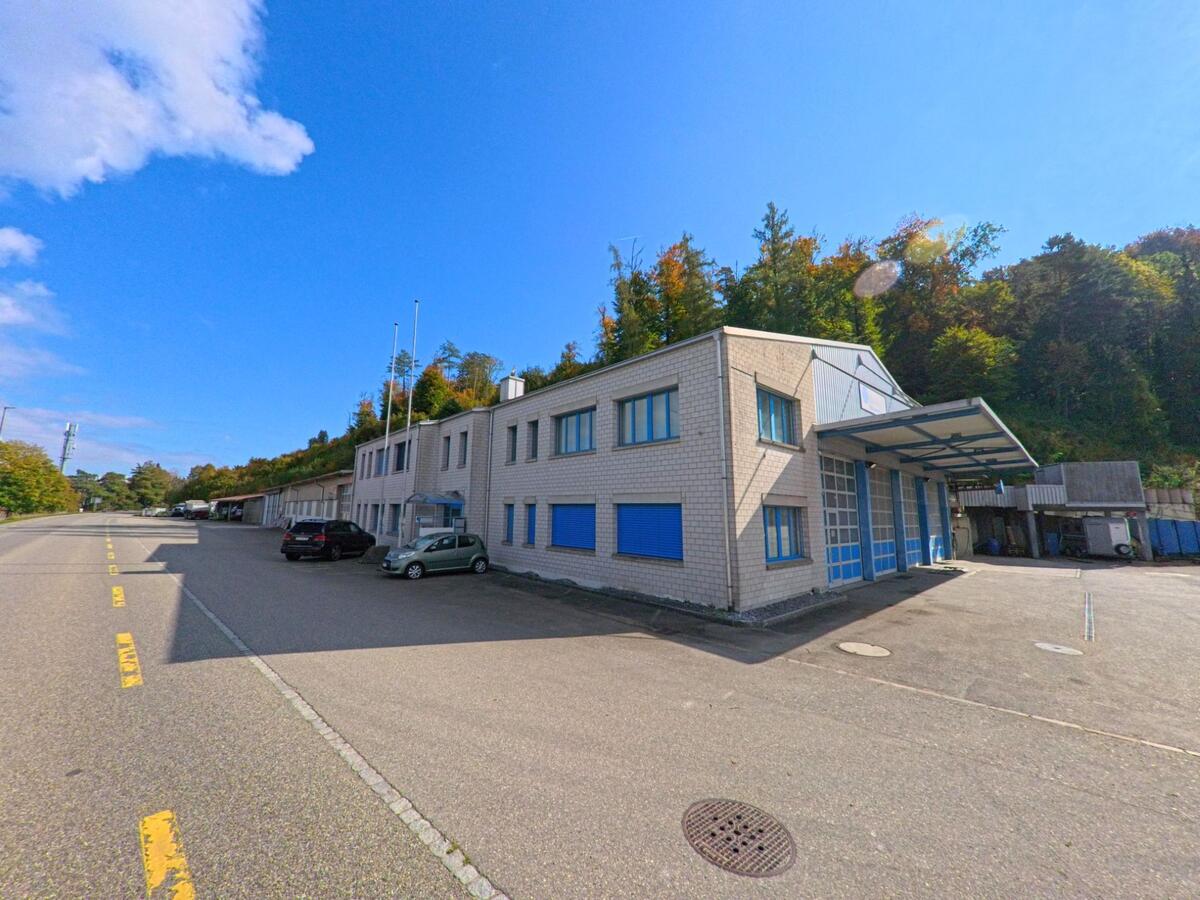 Ein modernes Industriegebaeude mit blauen Fensterlaeden und breiter Einfahrt steht in Schwaderloch (5326, Kanton Aargau) am Rand eines bewaldeten Hanges unter klarem Himmel.