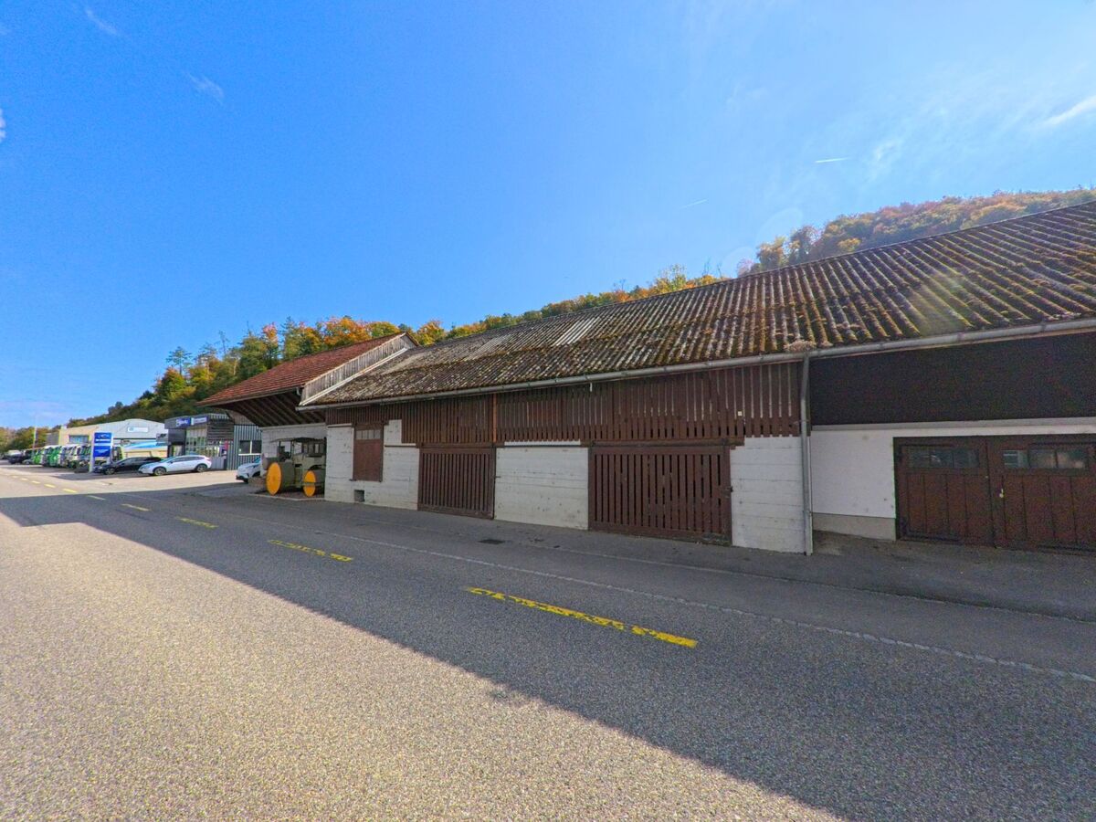 Eine langgezogene Lagerhalle mit Holzfassade und alten Dachziegeln steht an der Hauptstrasse in Schwaderloch (5326, Kanton Aargau) unter klarem Himmel am Rand eines bewaldeten Huegels.