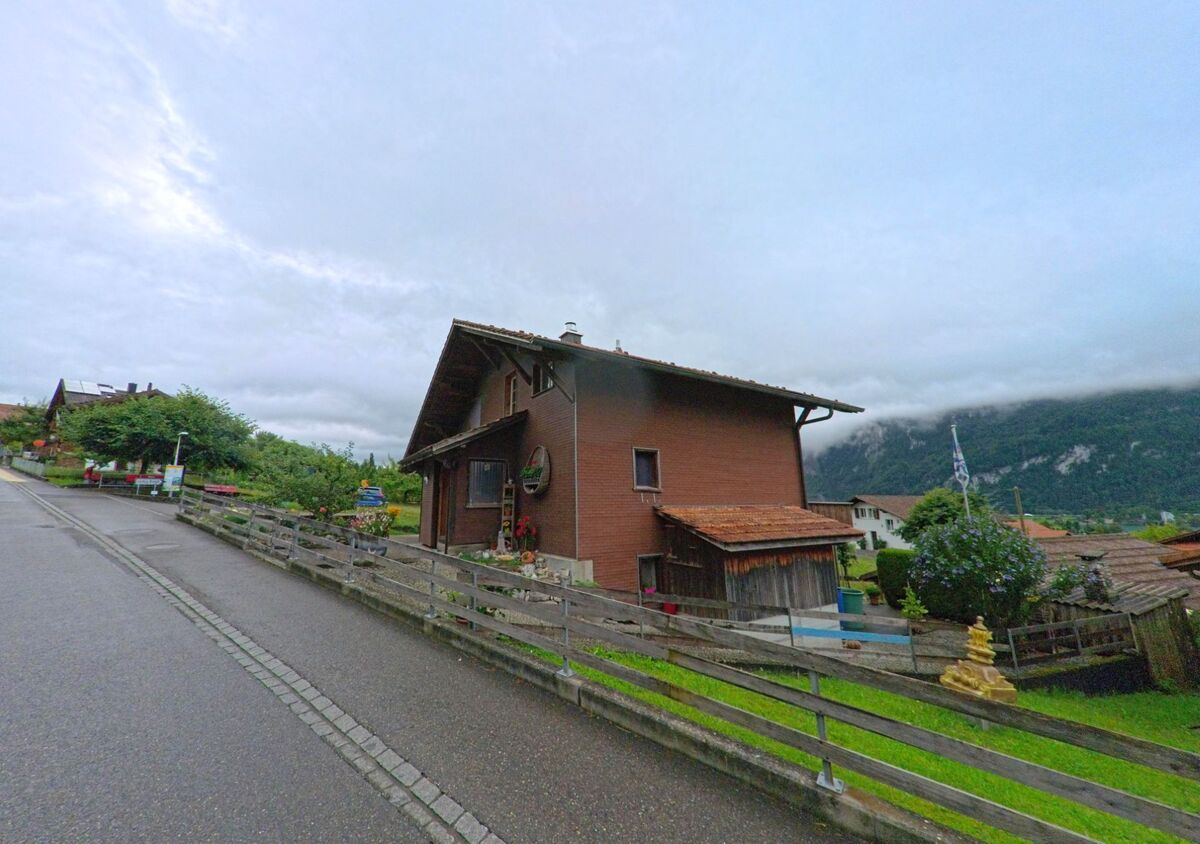 In Schwanden bei Brienz (3855, Kanton Bern) steht ein braunes Holzhaus mit Ziegeldach an einer leicht ansteigenden Strasse mit Blick auf die umliegenden Berge.