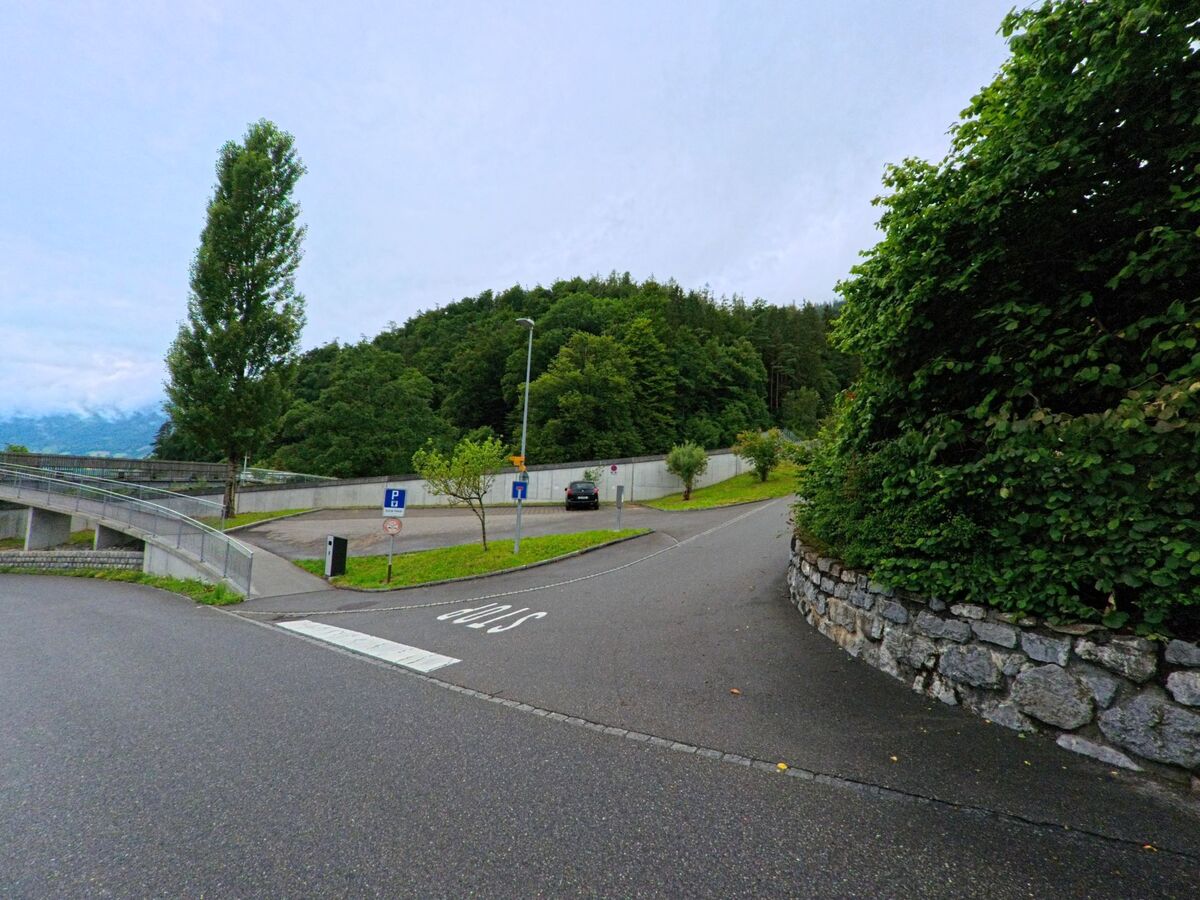 In Schwanden bei Brienz (3855, Kanton Bern) mündet eine Nebenstrasse mit Parkplatz in eine von Bäumen gesäumte Waldzufahrt, begrenzt durch eine niedrige Steinmauer.