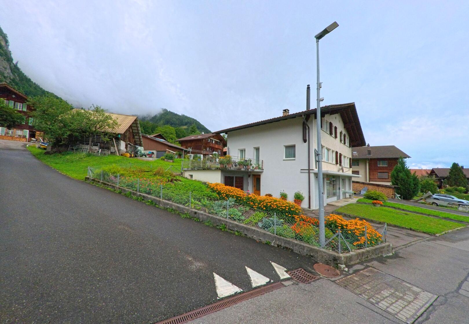 In Schwanden bei Brienz (3855, Kanton Bern) steht ein weisses Wohnhaus mit gepflegtem Vorgarten voller orangefarbener Blumen an einer Nebenstrasse.
