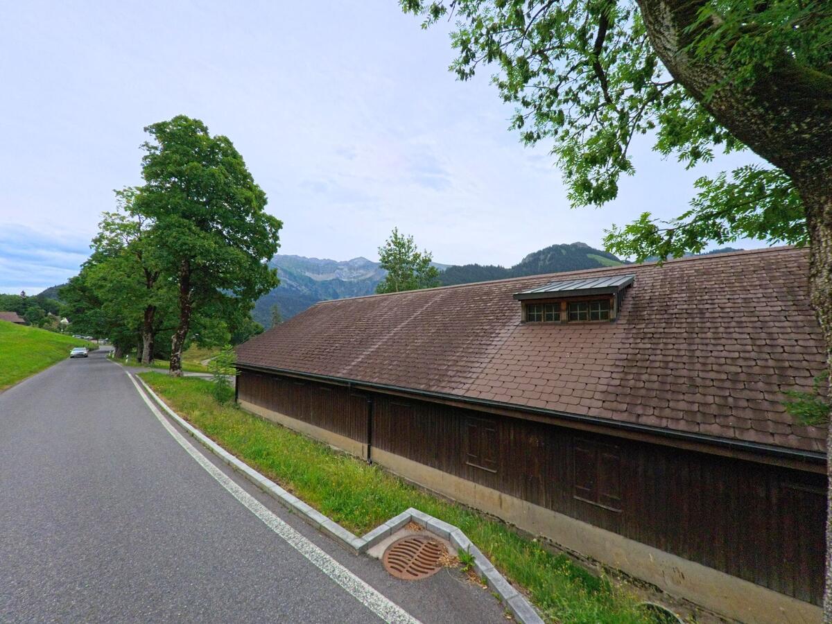 Eine schmale Strasse in Schwarzenberg (6013, Kanton Luzern) verläuft entlang eines langen Holzgebäudes mit Ziegeldach, eingerahmt von Bäumen und grünen Wiesen.