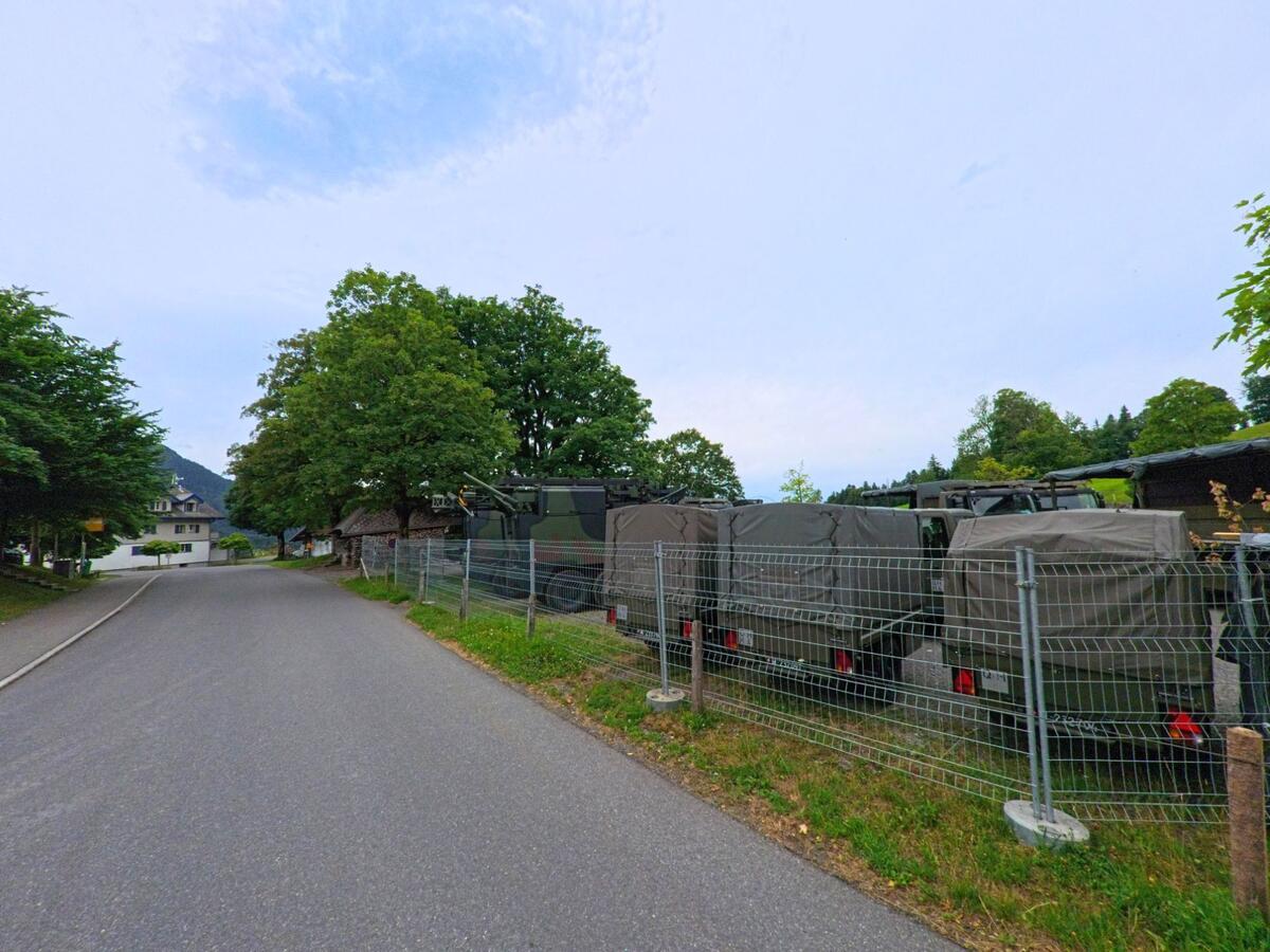 Entlang einer ruhigen Strasse in Schwarzenberg (6013, Kanton Luzern) sind mehrere Militärfahrzeuge hinter einem Metallzaun geparkt, flankiert von grossen Bäumen und Wiesen.