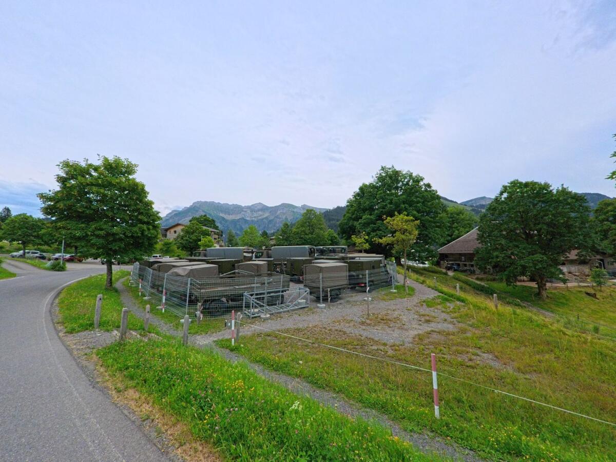 In Schwarzenberg (6013, Kanton Luzern) sind mehrere Militärfahrzeuge hinter einem Gitterzaun abgestellt, während sich im Hintergrund Wohnhäuser und bewaldete Hügel erstrecken.