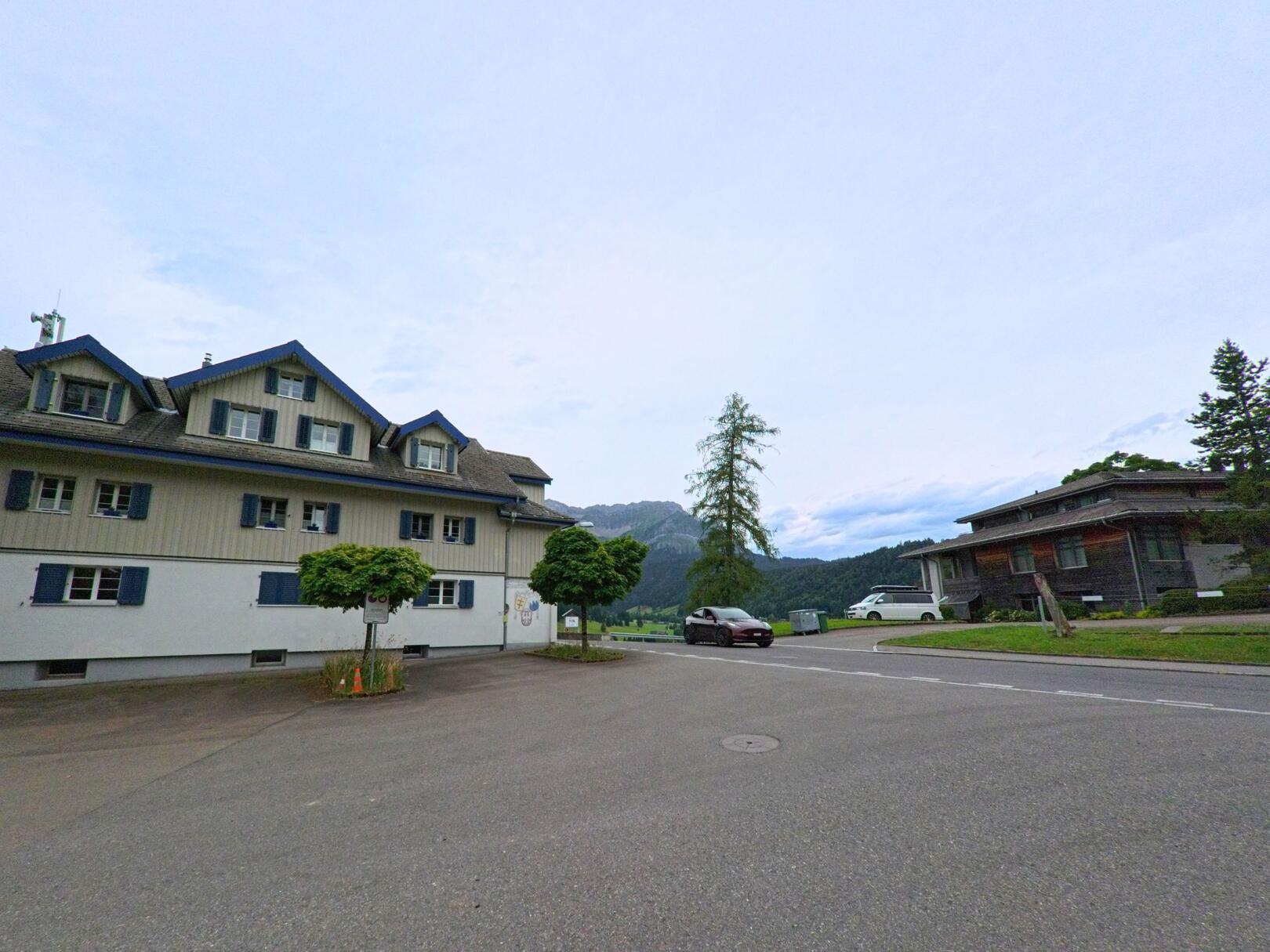In Schwarzenberg (6013, Kanton Luzern) stehen ein traditionelles Wohnhaus mit blauen Fensterläden und ein modernes Gebäude an einer ruhigen Strasse mit Blick auf bewaldete Hügel.