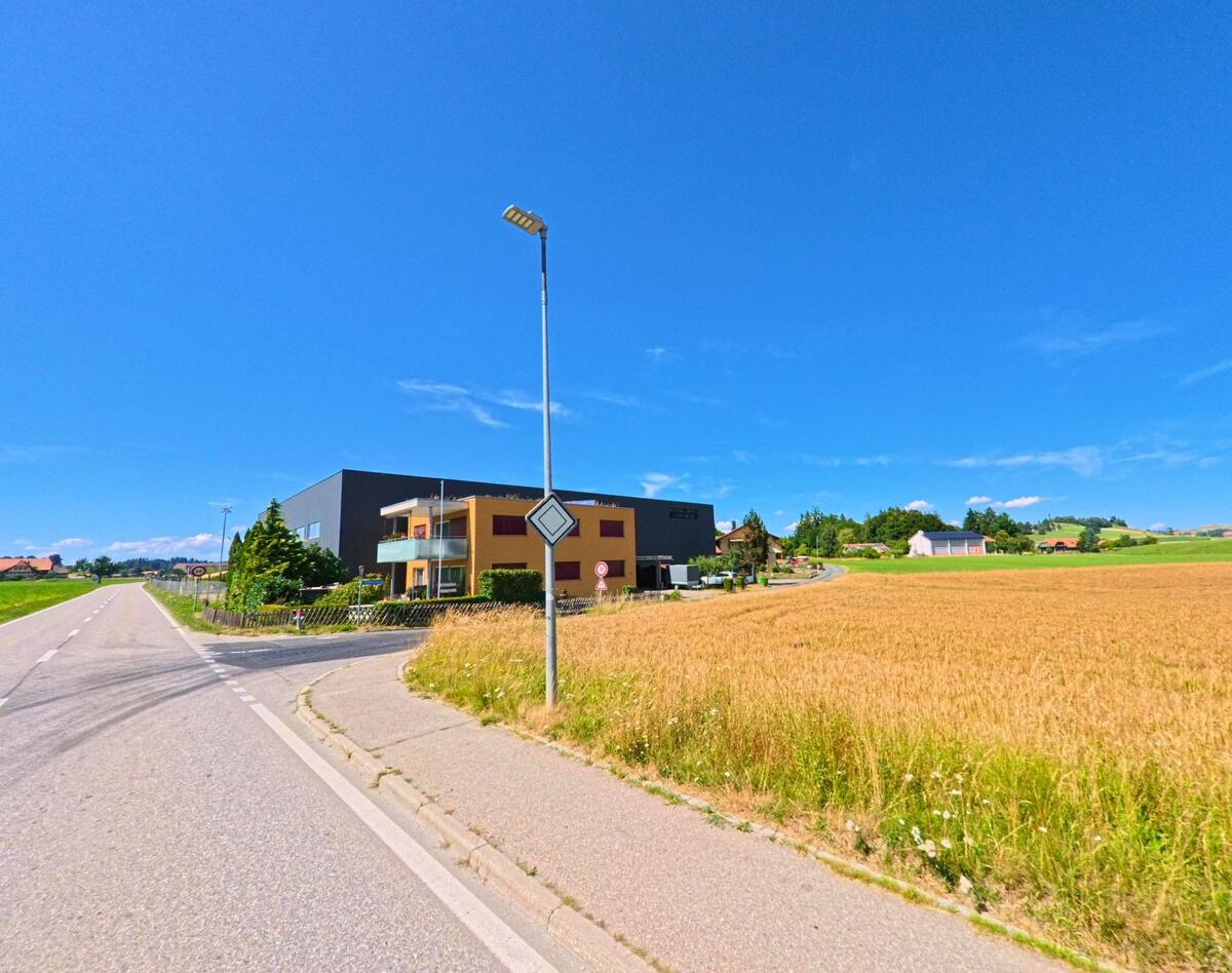 Am Rand eines Getreidefelds in Schwarzenburg (3152, Kanton Bern) liegt ein modernes Wohn- und Gewerbegebäude direkt an einer Landstrasse.