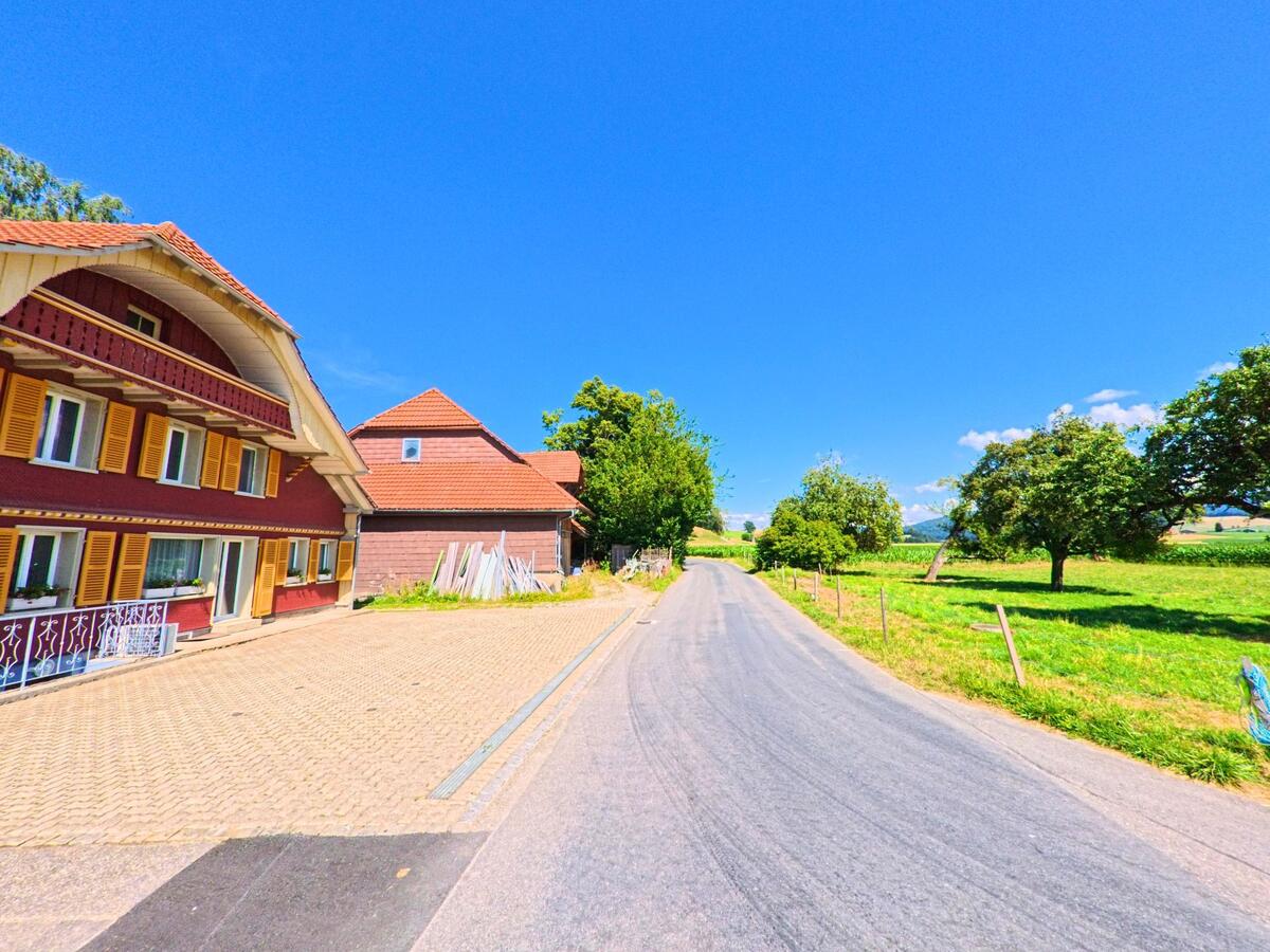 In Schwarzenburg (PLZ 3152, Kanton Bern) führt eine schmale Strasse vorbei an einem rot gestrichenen Wohnhaus mit gelben Fensterläden in eine grüne Wiesenlandschaft.