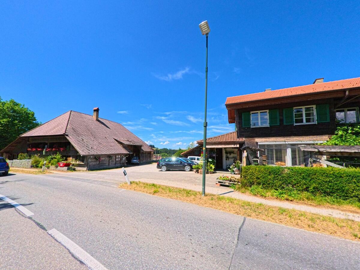 Entlang einer Hauptstrasse in Schwarzenburg (3152, Kanton Bern) stehen zwei ländliche Wohnhäuser mit gepflegten Vorgärten und parkenden Fahrzeugen.