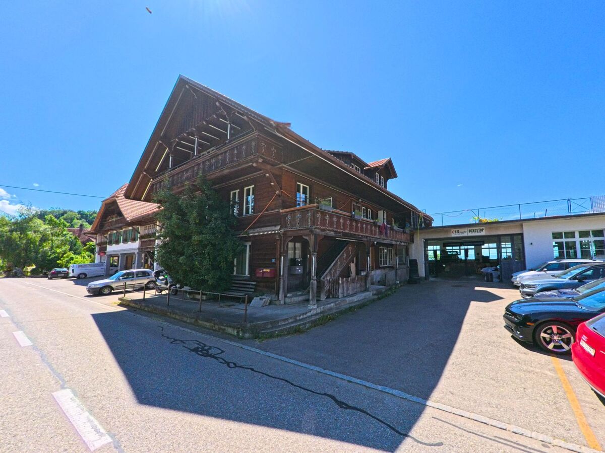 In Schwarzenburg (3157, Kanton Bern) steht ein historisches Holzhaus mit verziertem Balkon neben einem modernen Garagenbetrieb mit geparkten Autos.