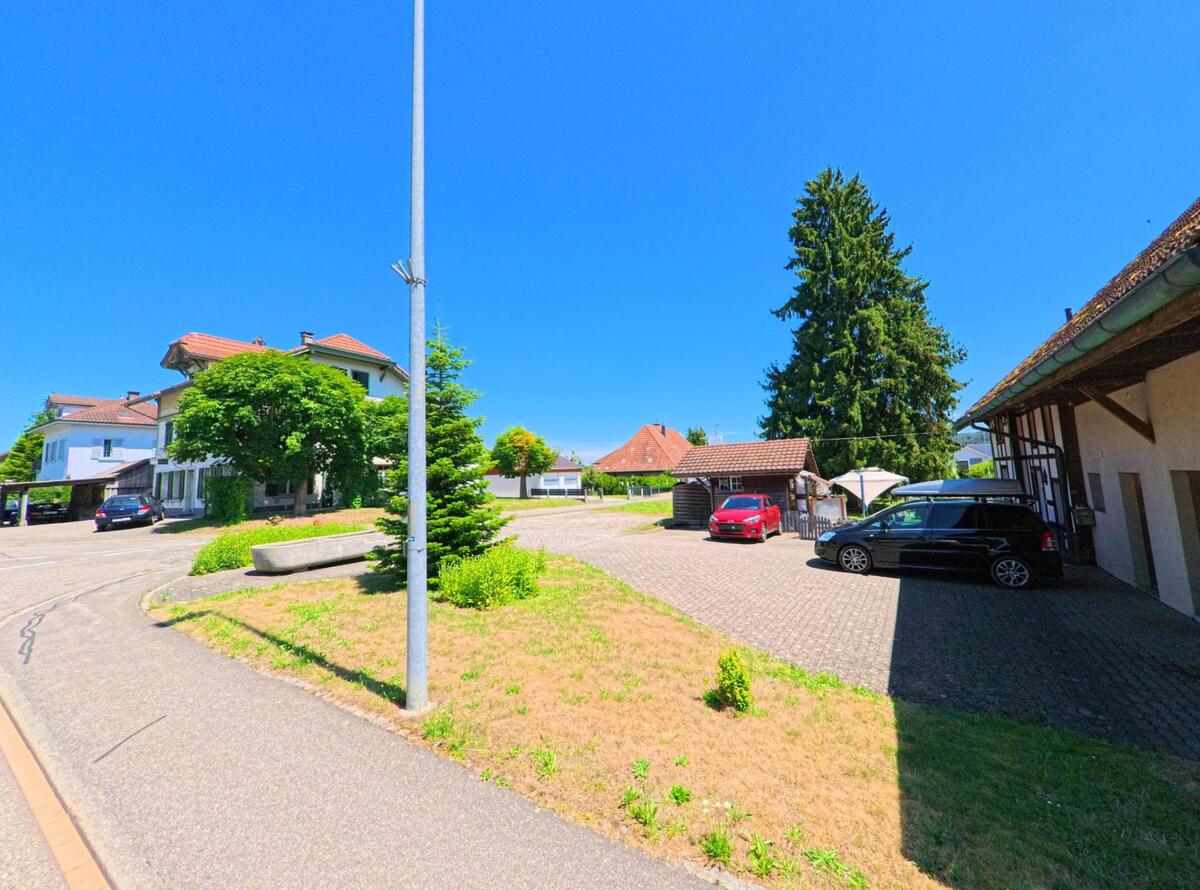 In Schwarzhäusern (4911, Kanton Bern) oeffnet sich zwischen Wohnhaeusern und einem Wirtschaftsgebaeude ein gepflasterter Platz mit mehreren geparkten Autos und schattenspendenden Baeumen
