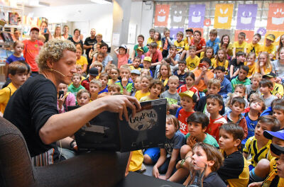 Ein Mann liest vor einer grossen Gruppe von Kindern aus einem Buch, waehrend sie aufmerksam zuhoeren; die Szene spielt sich in Zürich (8006, Kanton Zürich) in einem Veranstaltungsraum ab.