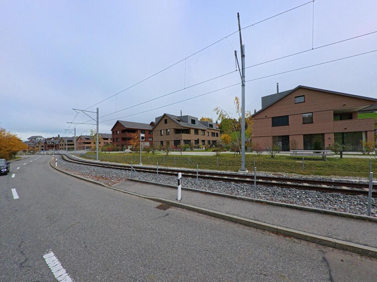 Entlang einer Strasse in Schwende (9057, Kanton Appenzell Innerrhoden) verläuft eine Bahnlinie mit Oberleitung neben modernen Wohnhäusern und begrünten Aussenanlagen.