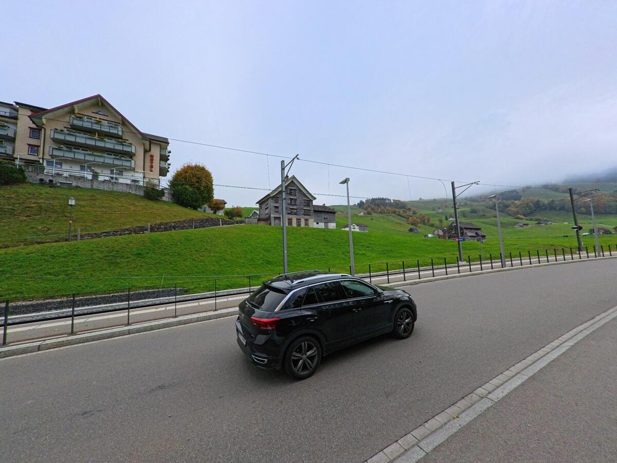 Entlang einer Strasse in Schwende (9057, Kanton Appenzell Innerrhoden) liegt eine Bahnstation mit Oberleitung vor grünen Hängen, einzelnen Häusern und nebelverhangenen Hügeln..jpg