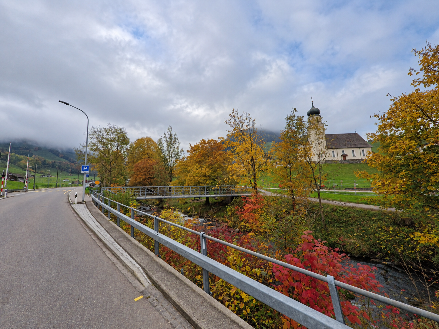 Entlang einer Strasse in Schwende (9057, Kanton Appenzell Innerrhoden) führt eine kleine Brücke über einen Bach, während eine Kirche zwischen herbstlichen Bäumen am grünen Hang steht.
