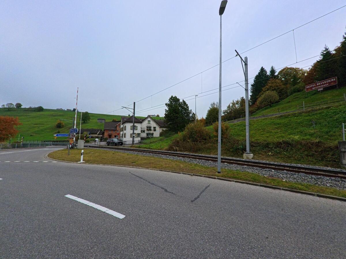An einer Strasse in Schwende (9057, Kanton Appenzell Innerrhoden) verläuft eine Bahnlinie mit Oberleitung neben grünen Hängen, Wohnhäusern und einem Bahnübergang..jpg